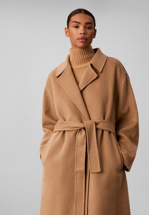 Femme portant un manteau en laine couleur camel ceinturé par-dessus un pull à col roulé côtelé assorti, debout devant un fond uni gris clair.