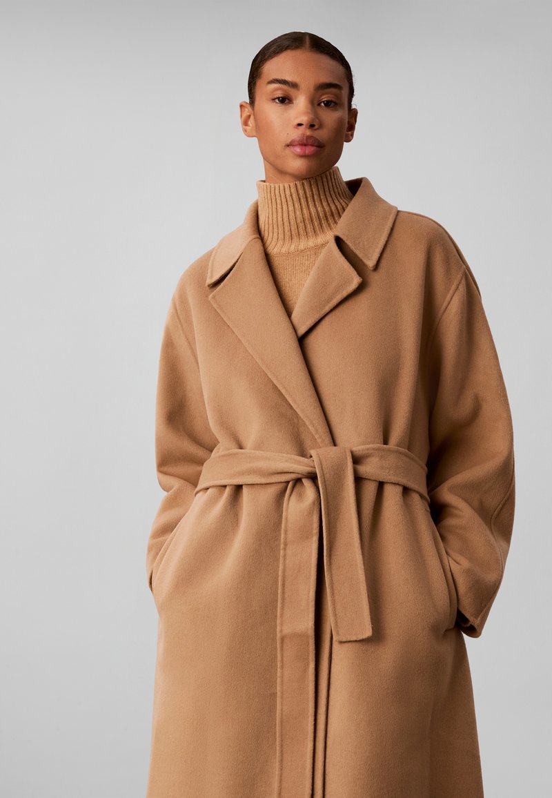 Femme portant un manteau en laine couleur camel ceinturé par-dessus un pull à col roulé côtelé assorti, debout devant un fond uni gris clair.