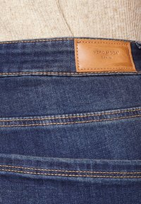 Traperice tamnoplave boje s kožnom oznakom u boji kože na pojasu koja glasi "VERO MODA". Imaju vidljivo šivanje i glatku teksturu.