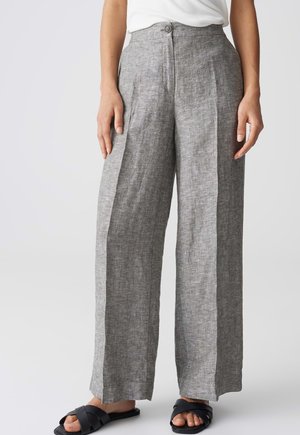 Frau trägt hoch taillierte, weit geschnittene graue Leinenhose mit vorderen Falten und schwarzen Slide-Sandalen, stehend vor einem weißen Hintergrund.