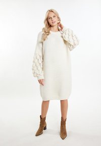 Abito maglione bianco lavorato a maglia con maniche a palloncino voluminose e texturizzate. Presenta polsini e orlo a coste, indossato con stivali alla caviglia marroni.