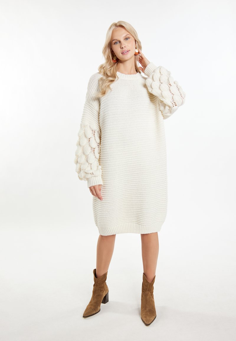 Abito maglione bianco lavorato a maglia con maniche a palloncino voluminose e texturizzate. Presenta polsini e orlo a coste, indossato con stivali alla caviglia marroni.