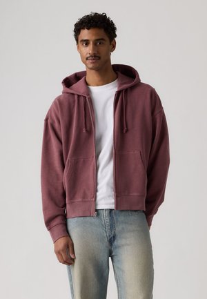 Mann trägt einen kastanienbraunen Kapuzenpullover mit Reißverschluss über einem weißen T-Shirt und ausgewaschenen blauen Jeans, steht vor einem schlichten hellen Hintergrund.