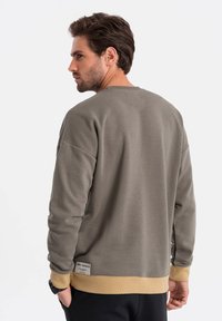 Grå sweatshirt med rund hals og lange ærmer. Har ribbede manchetter i beige og et lille mærke nederst til venstre. Blød tekstur.