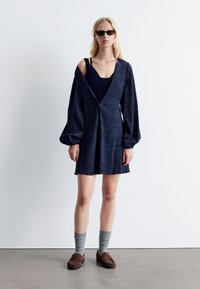 Navy-Kleid aus strukturiertem Stoff, tiefem V-Ausschnitt und Puffärmeln. Über einem figurbetonten navy-farbenen Oberteil getragen. Kombiniert mit braunen Loafers und grauen Socken.