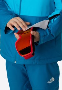 Ochelari de schi roșii, cu formă curbată și lentilă portocalie, sunt ținuți deasupra unei jachete albastre și a pantalonilor asortați. Materialul pare neted și durabil.