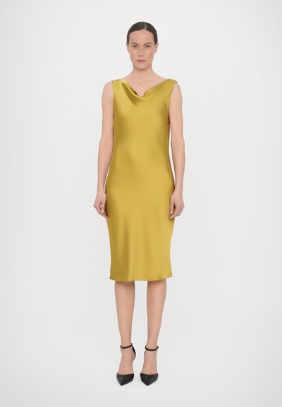 Satin-gelbes Kleid mit Wasserfall-Ausschnitt, schmaler Passform, knielang und ohne Ärmel. Getragen mit schwarzen spitzen Pumps.