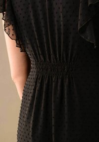Mustapuku, jossa on teksturoitu kangas ja pistekuvio. Takana on smokkityylinen yksityiskohta ja läpikuultavat röyhelösateet, jotka korostavat suunnittelua.