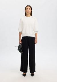 Hvit, teksturert genser med puffede ermer, kombinert med svarte bukser med vide ben. Modellen holder en liten svart håndveske.