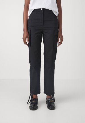 Zwarte cargo broek met een slimfit ontwerp, voorzien van zijzakken, een ritsgulp en enkelbinders, gemaakt van een stevige stof. Gecombineerd met zwarte schoenen.