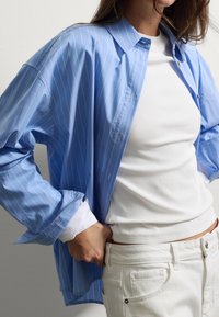 Mujer vistiendo una camisa azul con rayas verticales suelta sobre una camiseta de tirantes blanca y jeans blancos, con las manos metidas en los bolsillos frontales.