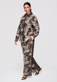 Femme debout en chemise et pantalon marron assortis avec un imprimé de feuilles blanches, portant des talons ouverts marron, main dans la poche.