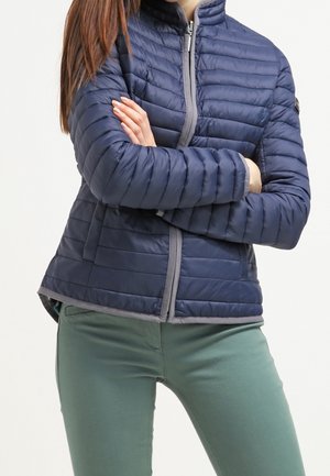 Navy gewatteerde jas met horizontale gestikte lijnen, grijze rits en kinbescherming, gecombineerd met lichtgroene strak zittende broek.