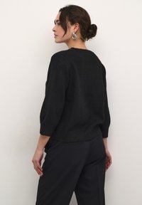 Pull en maille noir à manches trois-quarts, col rond et coupe décontractée, associé à un pantalon sombre bien ajusté.