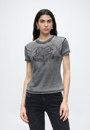 Femme aux longs cheveux foncés portant des boucles d’oreilles anneaux argentées, un t-shirt Diesel gris délavé et un jean noir sur un fond clair uni.