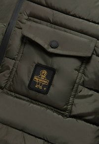 Tessuto di giacca a piumino verde oliva con texture trapuntata, caratterizzato da una tasca con patta e un logo patch nero con design di un cane giallo.