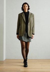Blazer verde oliva, blusa nera strutturata, gonna grigia, calze scure e sneakers nere. Vestibilità Slim, design strutturato con revers e tasche.