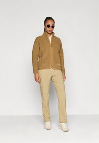 Bež flis jakna z zadrgo spredaj in stranskimi žepi, kombinirana s bež hlačami in svetlimi čevlji, ki jo nosi oseba z očali za sonce.