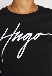 Henkilö, jolla on päällään musta pyöreäkaulus t-paita, jossa on isoilla valkoisilla kauniilla kirjaimilla "Hugo" kirjoitettuna rinnan kohdalla.