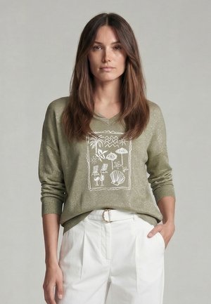 Frau mit langen braunen Haaren, die einen olivgrünen Pullover mit weißen Strandmotiven und weiße Hosen mit einer runden Gürtelschnalle trägt.