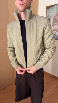 Šviesiai žalias bomberio stiliaus švarkas su aukštu apykakle, priekiniais užtrauktukais ir raištinėmis rankovėmis. Dėvimas ant juodos marškinėlių ir derinamas su tamsiomis šortais.