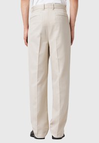 Beige pants made of cotton blend fabric, featuring a straight-leg design, double pleats, and two back pockets, worn with black footwear.