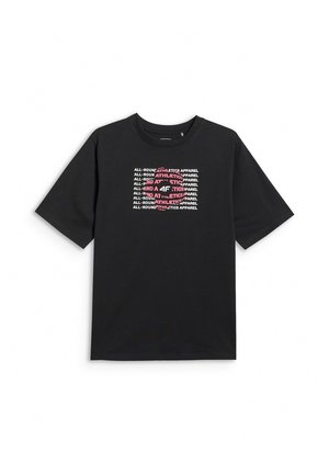 Młoda osoba stoi, ubrana w czarny oversize t-shirt z białym i czerwonym napisem oraz różowe spodnie na jednolitym szarym tle.