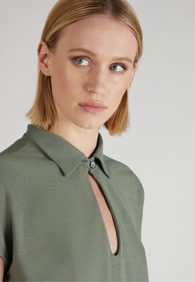 Blondynka z niebieskimi oczami, ubrana w oliwkową koszulę z kołnierzykiem i dekoltem w kształcie klucza, patrzy lekko na bok na jednolitym tle.