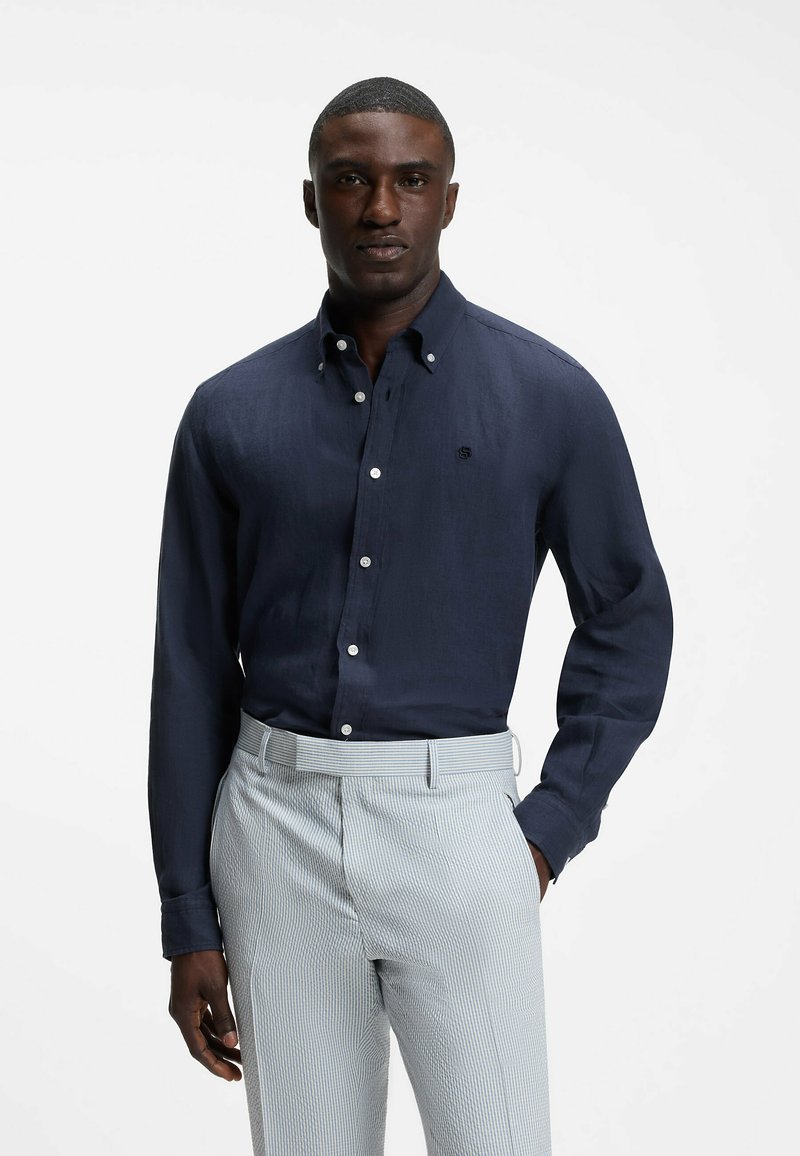 Homme portant une chemise bleu foncé boutonnée et un pantalon à rayures gris clair, debout avec une main dans la poche devant un fond uni.