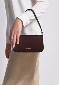 Borsa a pochette in pelle bordeaux con finitura testurizzata, logo dorato, tracolla sottile e bordi arrotondati, tenuta in mano contro uno sfondo chiaro.