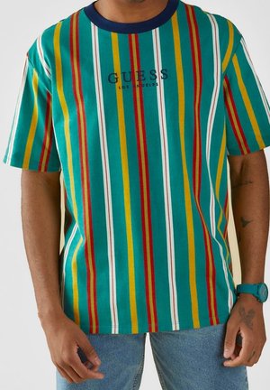 Man die een T-shirt met korte mouwen draagt met verticale strepen in turquoise, geel, rood en wit met de tekst "GUESS Los Angeles" en een blauwe spijkerbroek.