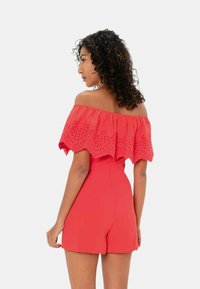 Romper rosso off-the-shoulder con dettagli in pizzo ricamato, corpetto aderente e pantaloni corti larghi. Texture liscia, senza alcun hardware visibile.
