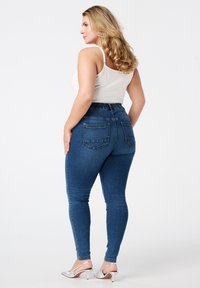 Un jean bleu taille haute avec une ceinture élastique, deux poches arrière, et une coupe slim, associé à un débardeur blanc et des talons argentés.
