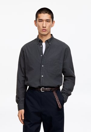 Mężczyzna ubrany w ciemnoszarą koszulę zapinaną na guziki, na białej podkoszulce, granatowe spodnie i pleciony brązowy pasek, stoi z jedną ręką w kieszeni.