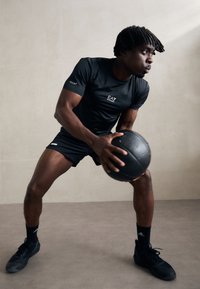 Svart atletisk t-skjorte og shorts, holder en svart medisinball. Har en logo på ermet til skjorten. Skoene er svarte med detaljer.
