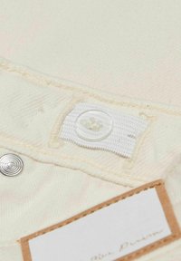 Beige tyg med en strukturerad yta, med en liten vit knapp och metalltryckknapp. En läderetikett är fäst med text.