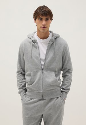 Jonge man draagt een lichtgrijze hoodie met rits en bijpassende joggingbroek, met een wit T-shirt, staat met handen in de zakken.