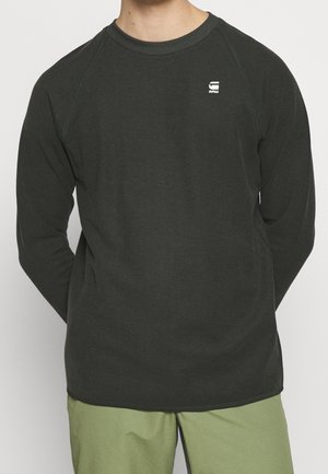 Mężczyzna ubrany w ciemną, teksturowaną koszulę z długim rękawem z małym białym logo na piersi oraz jasnooliwkowe spodnie, stoi z rękami za plecami.