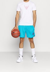 Hombre con camiseta blanca con logo de toro, pantalones cortos de baloncesto azules, calcetines blancos y zapatillas sosteniendo un balón de baloncesto a su lado.
