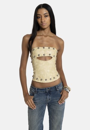 Kvinde med langt sort hår iført beige stropløs top med metalnitter og udskæring, blå jeans og metalliske armbånd, stående med hænderne nær lommerne.