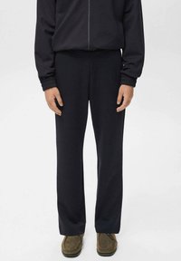 Persoon in zwarte rechte broek, zwarte zip-up jas met elastische manchetten en olijfgroene suède schoenen, staand op een witte achtergrond.