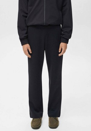 Persona con pantalones rectos negros, chaqueta negra con cremallera y puños elásticos, y zapatos de ante verde oliva, de pie sobre un fondo blanco.