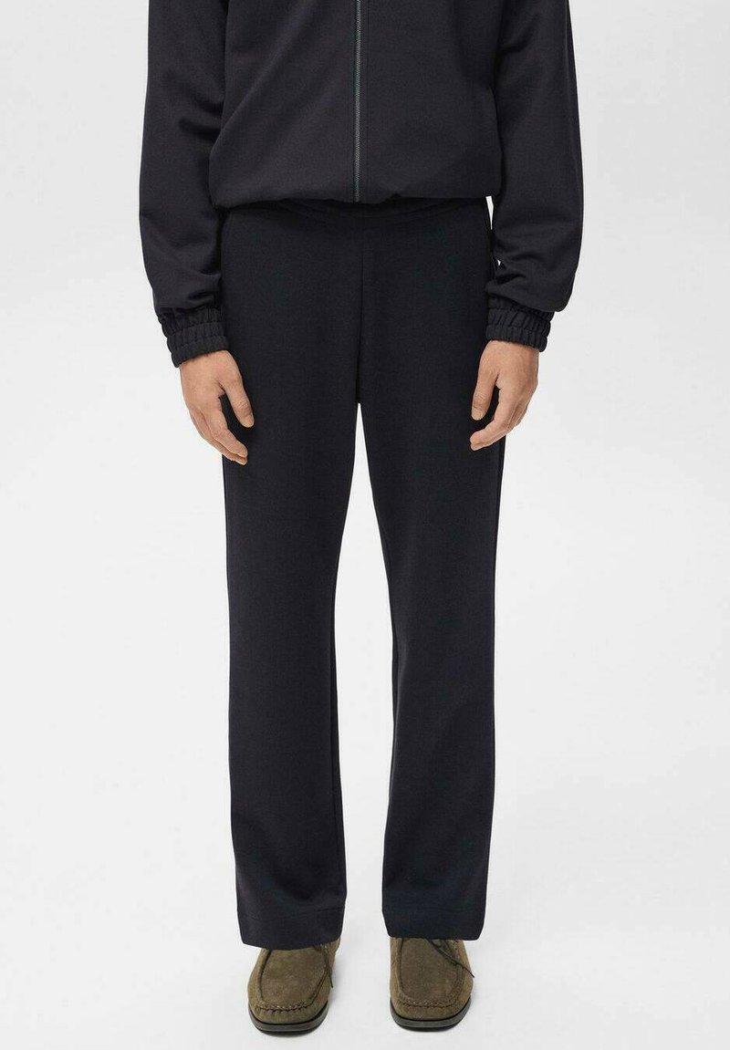 Persoon in zwarte rechte broek, zwarte zip-up jas met elastische manchetten en olijfgroene suède schoenen, staand op een witte achtergrond.