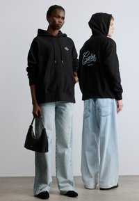 Čierna mikina s kapucňou s predným vreckom a striebornými šnúrkami, s bielym nápisom na chrbte; doplnená svetlomodrými širokými džínsami.