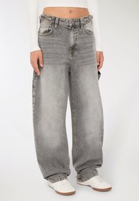 Osoba ubrana w luźne, szare, wyblakłe dżinsy z bocznymi kieszeniami i białe trampki, stojąca na jednolitym tle.