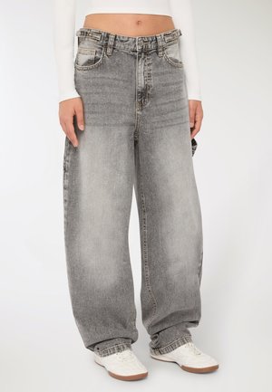Persona che indossa pantaloni grigi larghi e sbiaditi con tasche laterali e sneakers bianche, in piedi su uno sfondo semplice.
