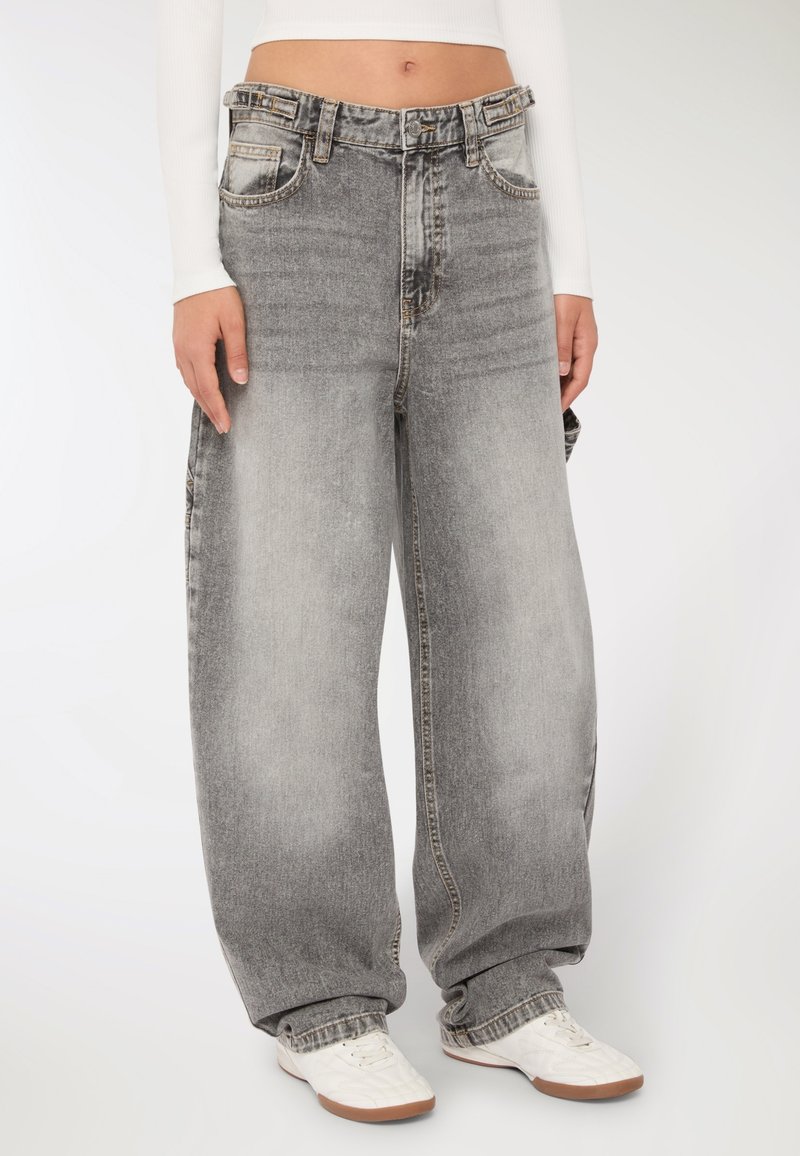 Osoba ubrana w luźne, szare, wyblakłe dżinsy z bocznymi kieszeniami i białe trampki, stojąca na jednolitym tle.