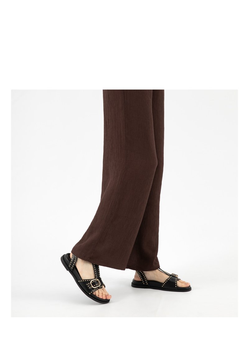 Person, die braune Hosen mit weitem Bein und schwarze Sandalen mit Nieten und Schnallen trägt, steht auf einer weißen Oberfläche.