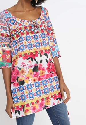 Femme portant une tunique colorée à motifs floraux et géométriques, associée à un jean bleu, sur un fond blanc uni.