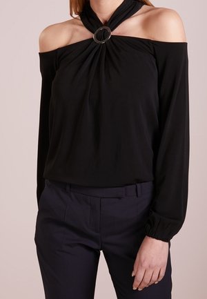 Kobieta ubrana w czarną bluzkę z odkrytymi ramionami, z detalem kołnierzyka na szyi oraz długimi rękawami, w połączeniu z ciemnymi, dopasowanymi spodniami.